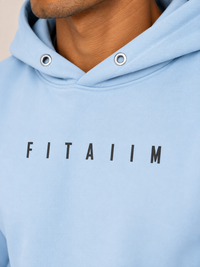 Close-up of a person wearing a light blue hoodie with the word 'FITAIIM' printed in black across the chest. The image shows the neck, chin, and upper chest of the model, with the hoodie hood and metal eyelets visible.