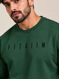 A close-up of a man wearing a dark green crewneck sweatshirt with the word 'FITAIIM' printed in black across the chest, set against a plain beige background.