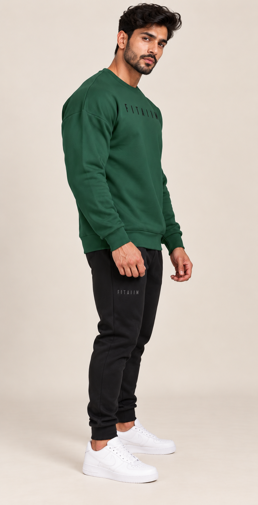 A man modeling a dark green crewneck sweatshirt and black joggers with white sneakers, standing against a solid beige studio background.