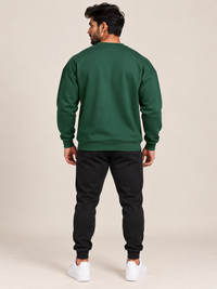 A man shown from the back wearing a plain green crewneck sweatshirt and black jogger, standing against a solid beige studio background.