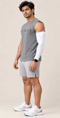 A man modeling a gray sleeveless athletic tank top with 'FITTAIIM' printed on the chest, paired with light gray shorts and white sneakers, wearing a white compression sleeve on one arm.