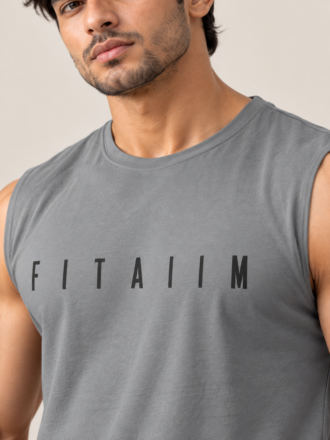 A close-up of a man wearing a gray sleeveless muscle tank top with the word 'FITAIIM' printed in black across the chest, set against a plain beige background.
