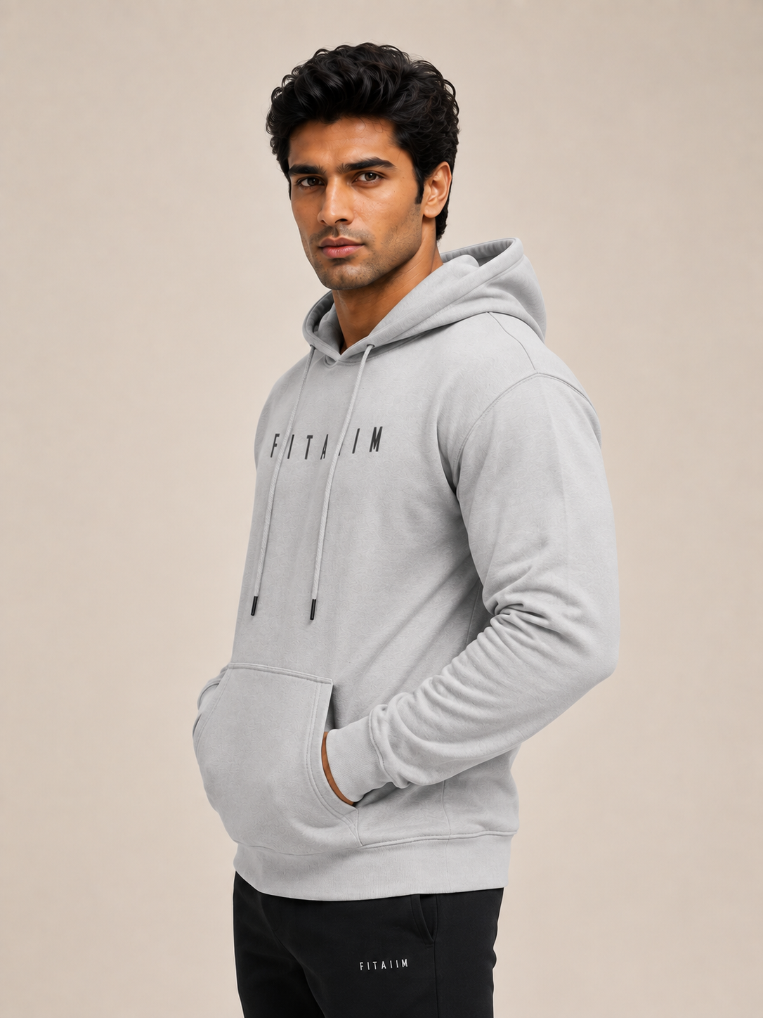 A man wearing a light gray fitted pullover hoodie with black drawstrings and black 'FITAIIM' lettering on the chest, paired with black pants, against a plain beige studio background.
