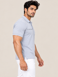 A man wearing a light gray heathered short-sleeve polo shirt with 'FITAIIM' printed on the chest, paired with white trousers, set against a solid beige background.