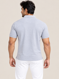 A man viewed from the back wearing a light gray short-sleeve polo shirt and white pants, standing against a plain beige studio background.