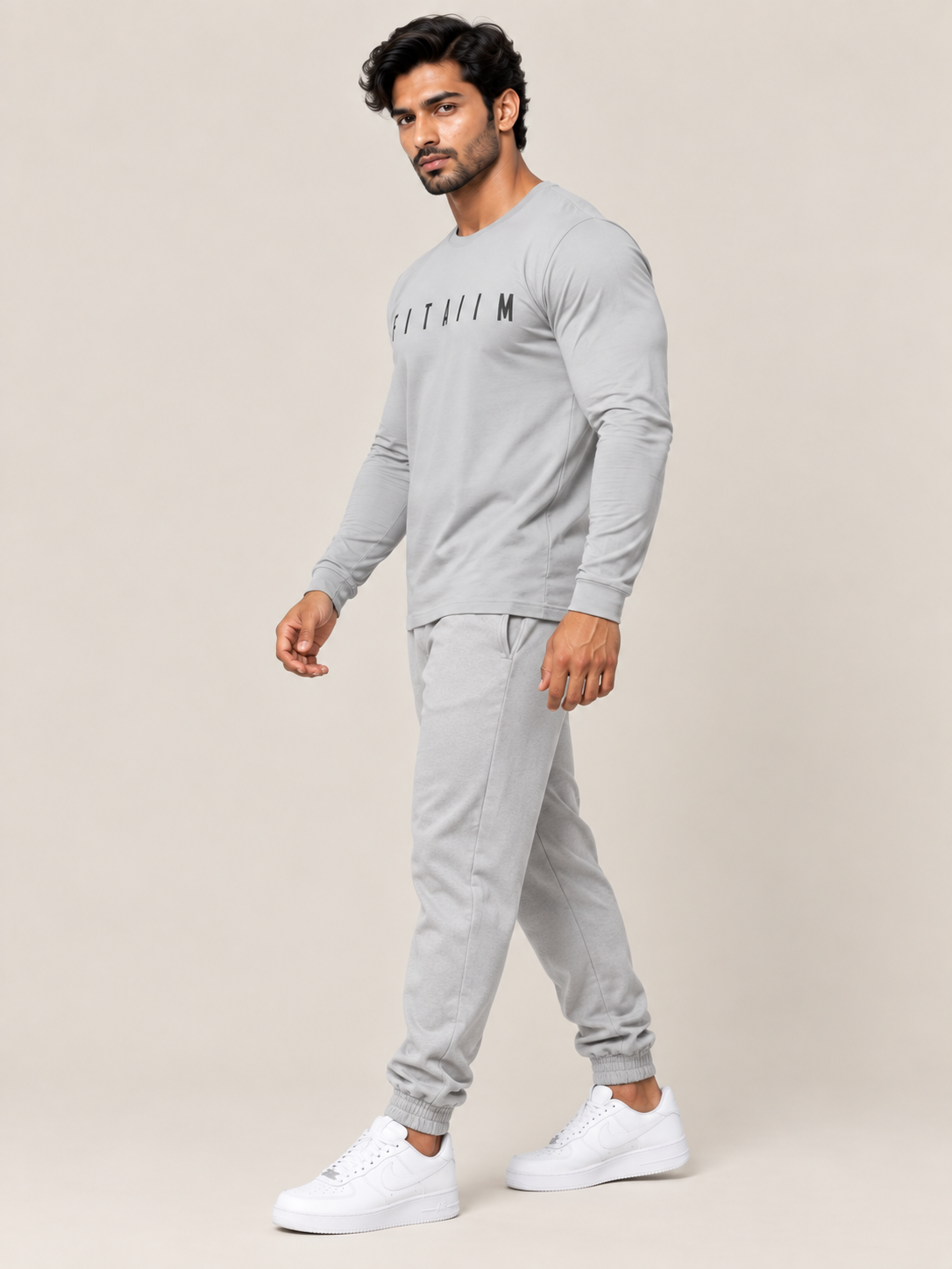 A man modeling a light gray long-sleeve crewneck shirt and matching jogger pants set, paired with white sneakers, against a solid beige studio background.