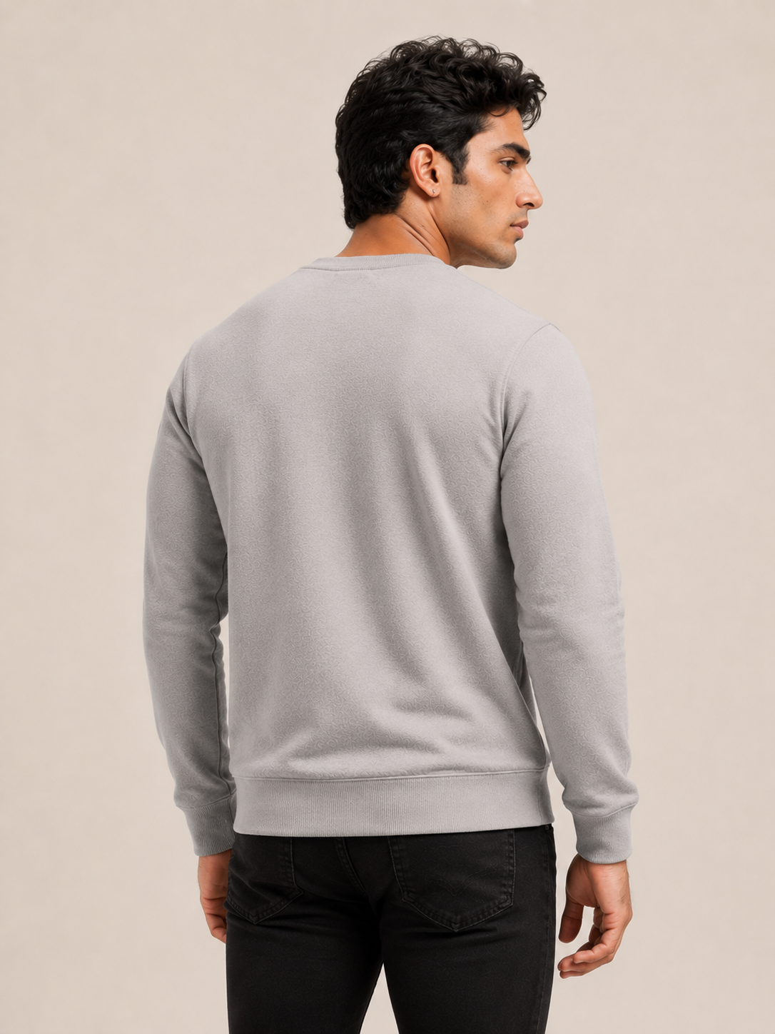A man viewed from the back wearing a grey long-sleeve crewneck sweater and light beige pants, set against a plain beige studio background.