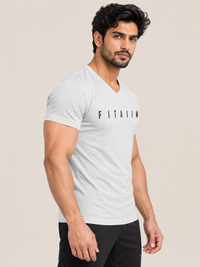 A muscular man wearing a light gray v-neck t-shirt with the brand name 'FITAIIM' printed in black across the chest, paired with black pants, set against a solid beige background.