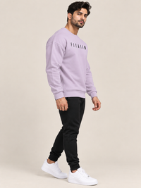 A man modeling a lavender crewneck sweatshirt with black text on the chest, paired with black joggers and white sneakers, against a solid beige background.