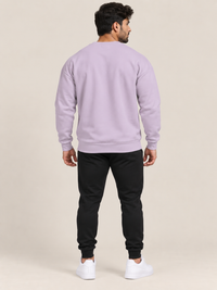 A man viewed from behind wearing a lavender crewneck sweatshirt, black jogger pants, and white sneakers, standing against a solid beige studio background.