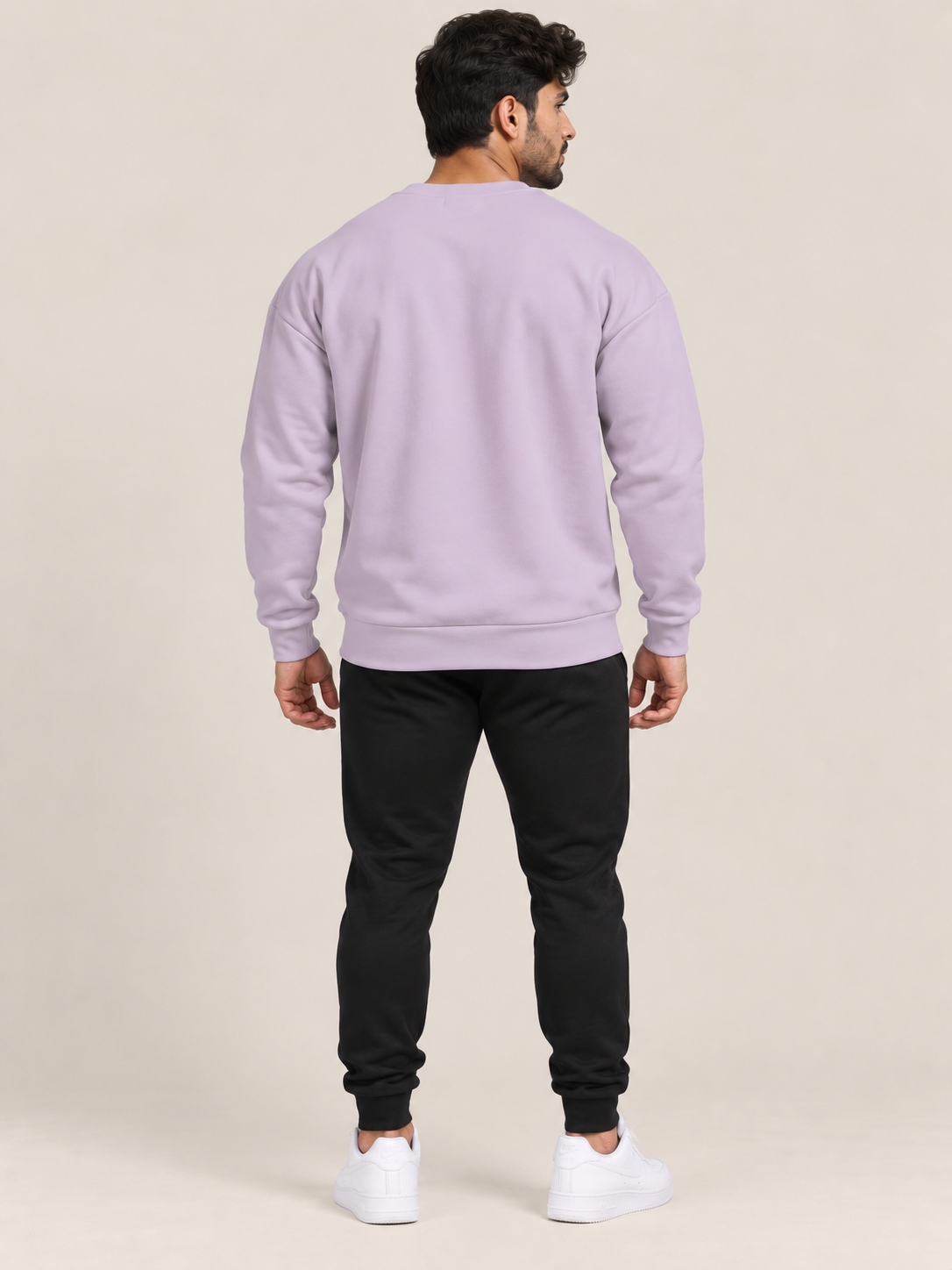 A man viewed from behind wearing a lavender crewneck sweatshirt, black jogger pants, and white sneakers, standing against a solid beige studio background.