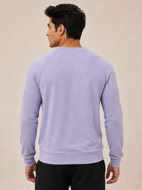 A man viewed from the back wearing a light lavender crewneck sweatshirt and black pants, set against a plain beige studio background.