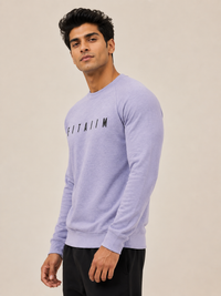 A man wearing a lavender sweatshirt  with a black graphic print on the chest, posed against a solid beige studio background.