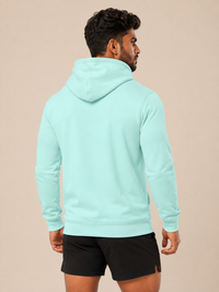 A man wearing a mint green pullover hoodie and black athletic shorts, shown from the back against a solid beige studio background.