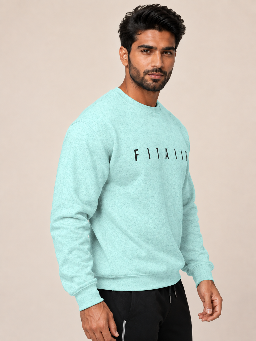 A man wearing a light mint green heather crewneck sweatshirt with 'FITALY' printed in black across the chest, paired with black pants, set against a solid beige background.
