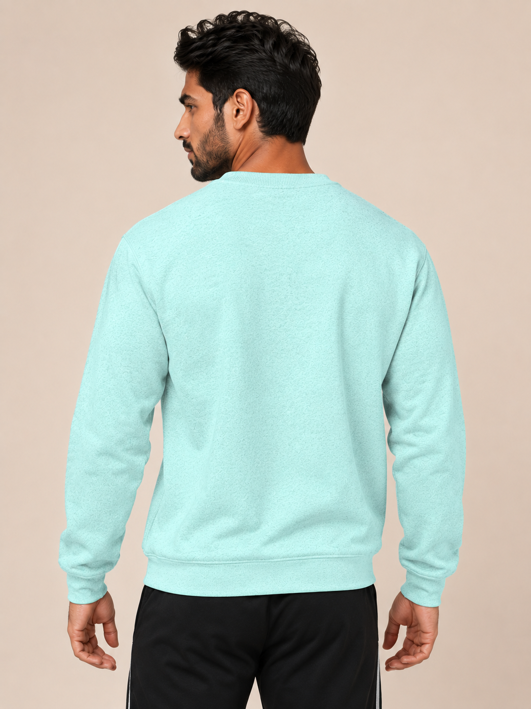 A man shown from the back wearing a light mint green crewneck sweatshirt and black track pants, set against a solid beige studio background.