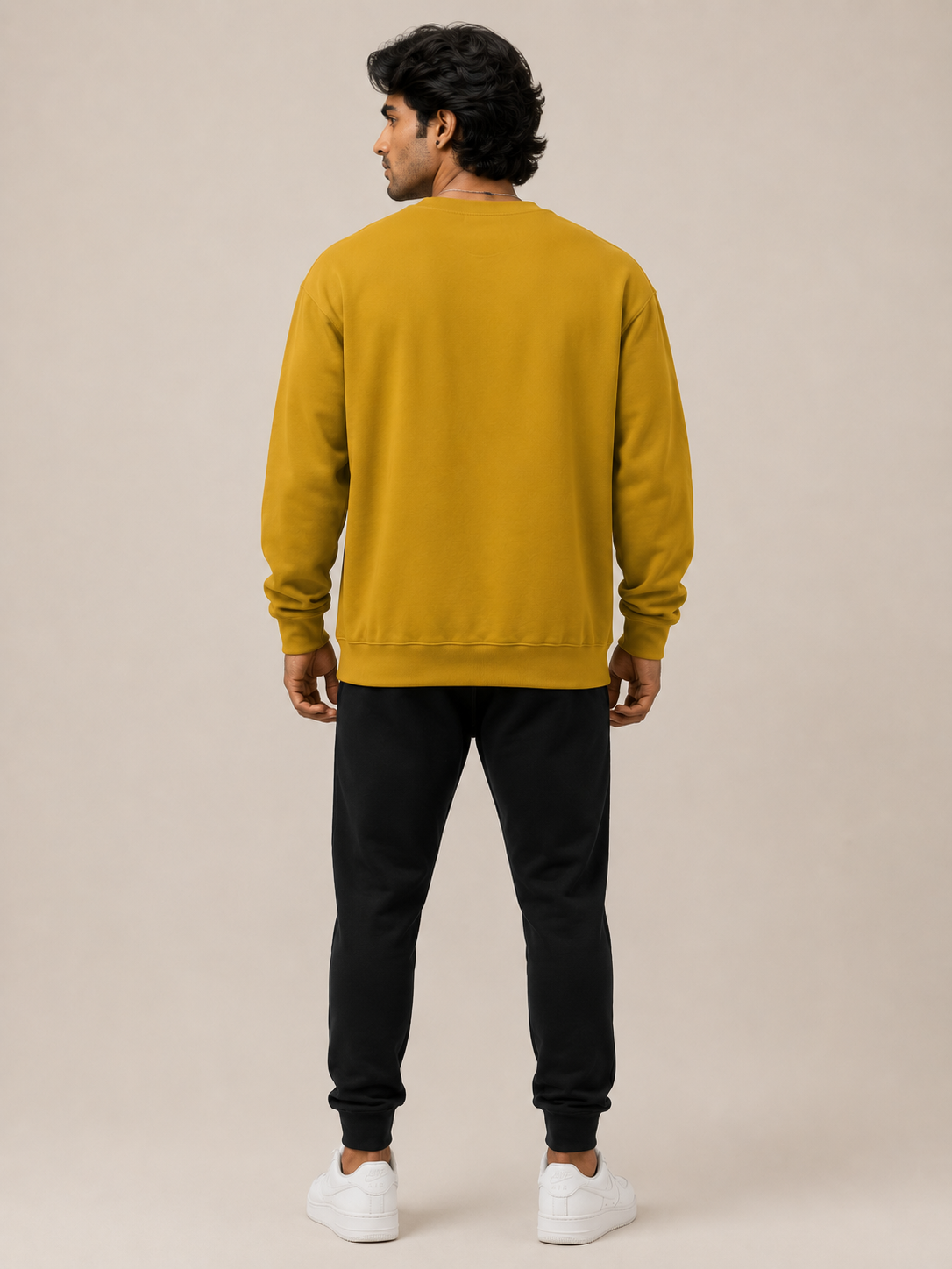 A man viewed from behind wearing a mustard yellow crewneck sweatshirt, black jogger pants, and white sneakers, standing against a solid beige studio background.