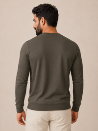 A man viewed from the back wearing a dark olive green long-sleeve crewneck sweater and light beige pants, set against a plain beige studio background.