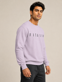A man wearing a light PINK long-sleeve crewneck sweatshirt with 'FITAIM' printed across the chest, paired with dark gray sweatpants, against a solid beige background.