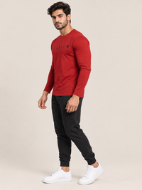 A man modeling a red long-sleeve crewneck t-shirt with black lettering on the chest, paired with black joggers and white sneakers, against a solid beige studio background.