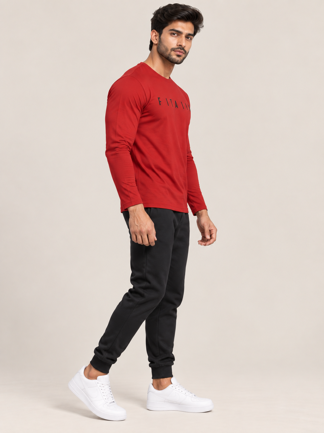 A man modeling a red long-sleeve crewneck shirt with black joggers and white sneakers, standing against a solid beige studio background.
