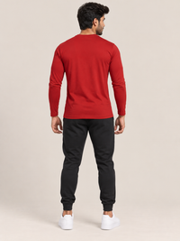 A man viewed from the back wearing a red long-sleeve crewneck shirt, black jogger pants, and white sneakers, standing against a plain beige studio background.