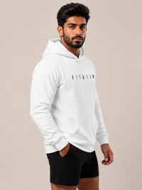 A man wearing a white pullover hoodie with 'FITAIIM' printed in black across the chest, paired with black shorts, standing against a solid beige studio background.