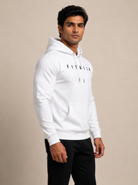 A man wearing a white pullover hoodie with 'FITAIIM' printed in black across the chest, paired with black pants, standing against a solid beige studio background.