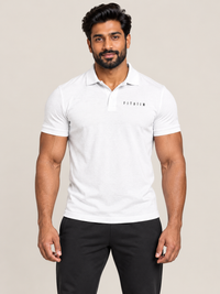 A muscular man wearing a white short-sleeve polo shirt with 'FItaiim' printed on the chest, paired with black pants, against a solid light beige background.