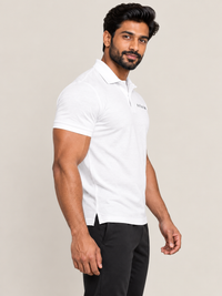A man wearing a white short-sleeve polo shirt with a small black embroidered logo on the left chest, paired with black trousers, photographed against a plain light beige studio background.