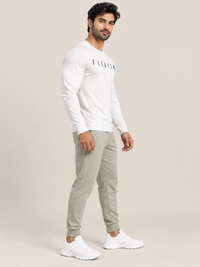 A man modeling a white long-sleeve crewneck sweatshirt with a black 'FITAIIM' logo on the chest, paired with light gray jogger pants and white sneakers, against a solid beige studio background.
