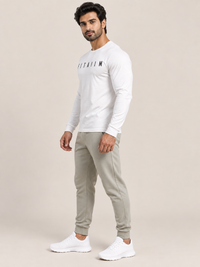 A man modeling a white long-sleeve athletic shirt with black 'FITAIIM' text on the chest, paired with light gray jogger pants and white sneakers, against a solid beige studio background.