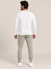 A man shown from the back wearing a white long-sleeve crewneck t-shirt, light beige jogger pants, and white sneakers, standing against a solid light beige studio background.