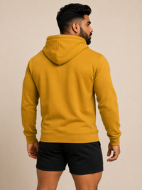 A man wearing a mustard yellow pullover hoodie, shown from the back, paired with black athletic shorts against a solid beige studio background.