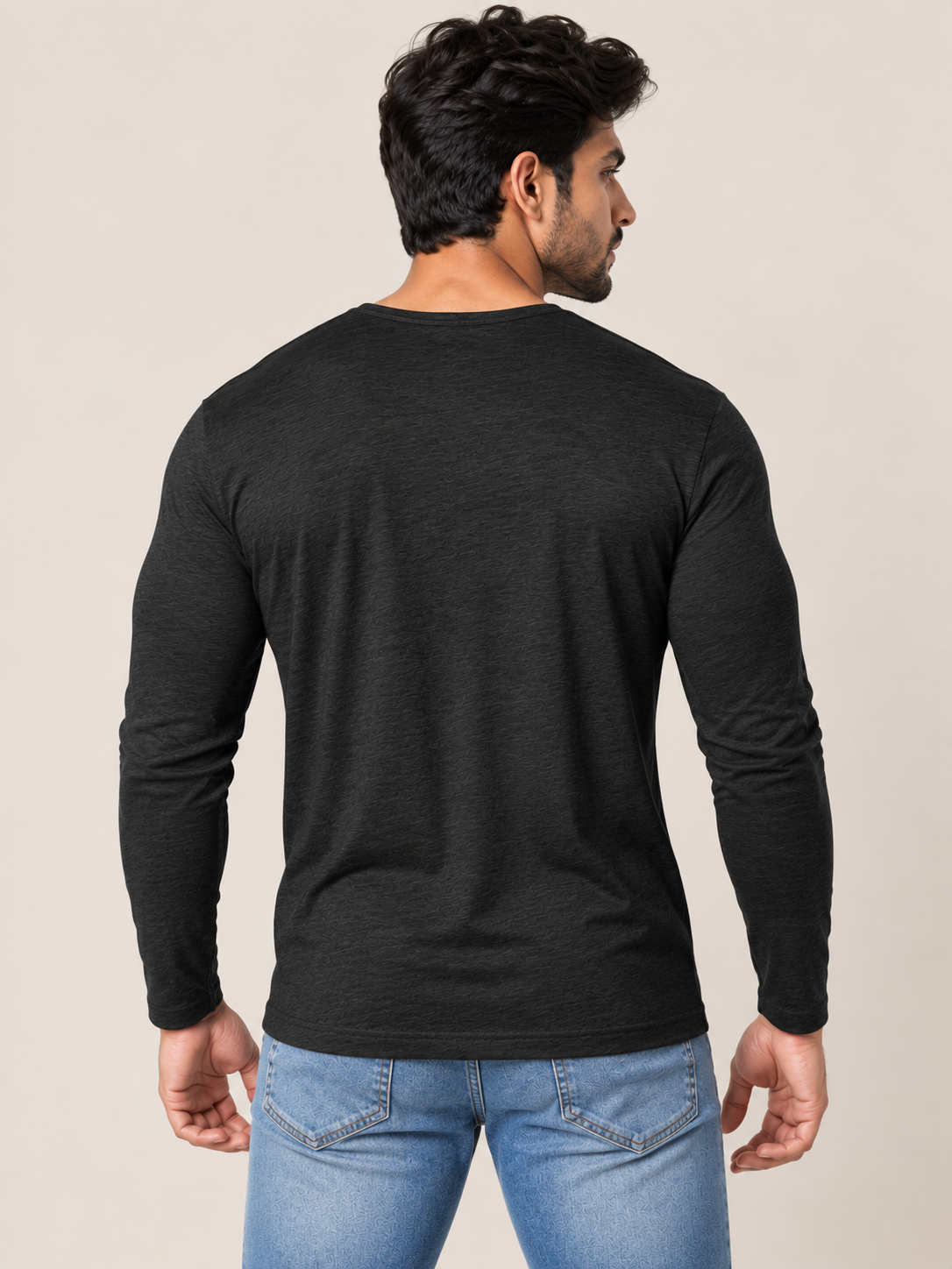 A man wearing a black long-sleeve crewneck t-shirt and blue jeans, shown from a rear three-quarter view against a solid light beige background.
