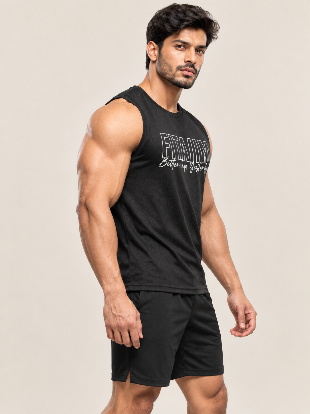 A muscular man wearing a black sleeveless athletic tank top with white text and black shorts, standing against a plain light beige studio background.