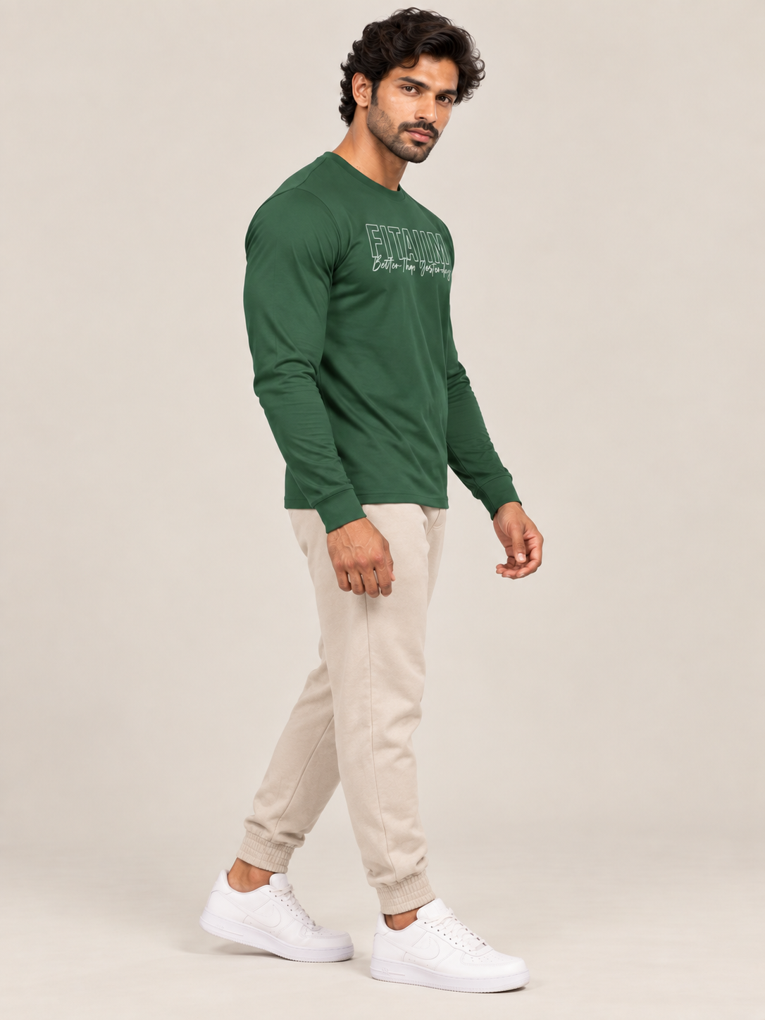 A man modeling a dark green long-sleeve crewneck shirt with a white graphic print on the chest, paired with beige jogger pants and white sneakers, against a solid light beige background.