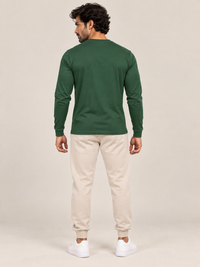 A man shown from the back wearing a dark green long-sleeve crewneck shirt, beige jogger pants, and white sneakers, standing against a plain light beige studio background.