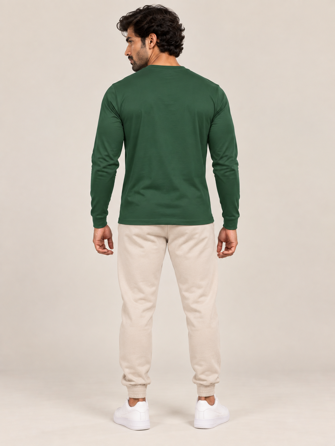 A man shown from the back wearing a dark green long-sleeve crewneck shirt, beige jogger pants, and white sneakers, standing against a plain light beige studio background.