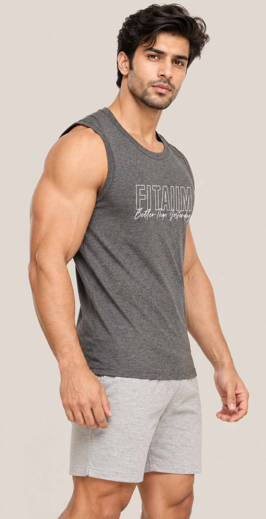 A muscular man wearing a dark gray sleeveless athletic tank top with 'FITAIIM Better Then Yesterday' printed on the front, paired with light gray shorts, against a solid beige background.