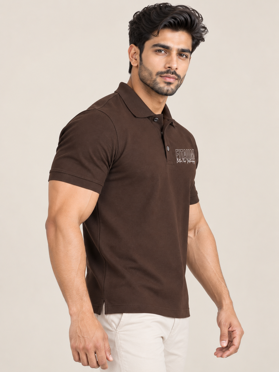 A man wearing a dark brown short-sleeve polo shirt with a small white embroidered logo on the left chest, paired with off-white trousers, set against a plain beige studio background.