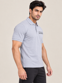 A man wearing a grey short-sleeve polo shirt with a small black logo on the left chest, paired with black pants, photographed against a plain light beige studio background.
