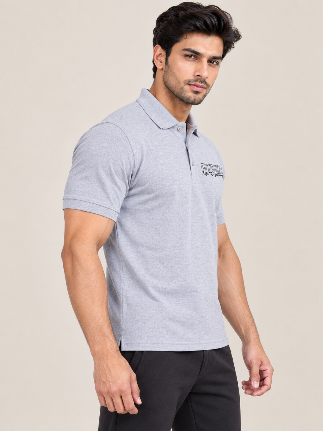A man wearing a grey short-sleeve polo shirt with a small black logo on the left chest, paired with black pants, photographed against a plain light beige studio background.