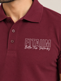 A close-up of a man wearing a maroon short-sleeve polo shirt with a white embroidered graphic on the chest that reads 'FITAIM Better Than Yesterday'.