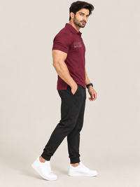 A man modeling a short-sleeve polo shirt in maroon, paired with black jogger pants and white sneakers, standing against a solid light beige background.