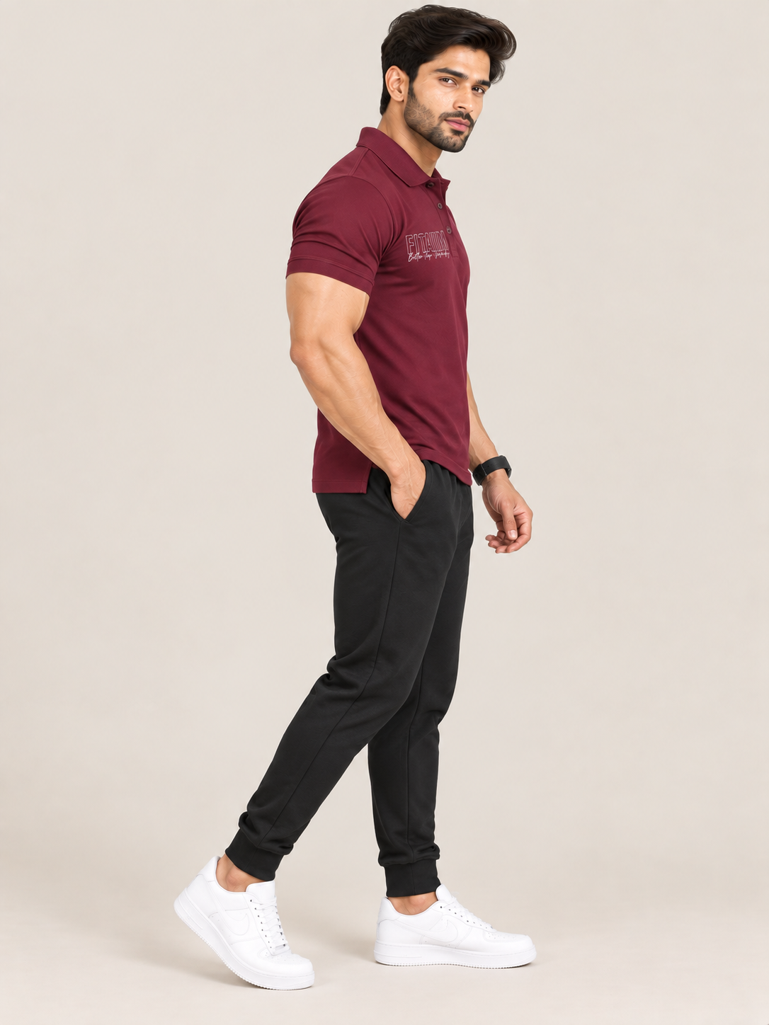 A man modeling a short-sleeve polo shirt in maroon, paired with black jogger pants and white sneakers, standing against a solid light beige background.