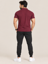 A man shown from the back wearing a short-sleeve polo shirt in maroon, black jogger pants, and white sneakers, standing against a plain beige studio background.