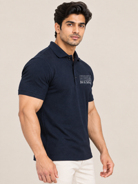 A muscular man wearing a dark navy blue polo shirt with a white embroidered logo on the left chest, paired with off-white trousers, set against a plain beige studio background.