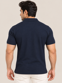 A man viewed from the back wearing a navy blue short-sleeve polo shirt and off-white trousers, set against a plain beige studio background.