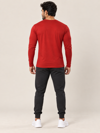 A man viewed from the back wearing a red long-sleeve crewneck shirt, black jogger pants, and white sneakers against a plain beige studio background.